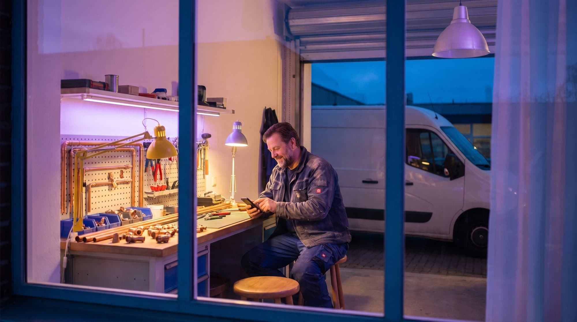 Installateur kijkt op zijn telefoon in de werkplaats, zijn witte bus buiten in de schemering